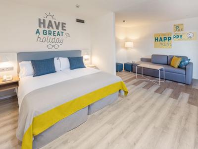 Helles Schlafzimmer mit großem Bett, blauen Kissen, gelben Decken und Sofa in modernem Design.