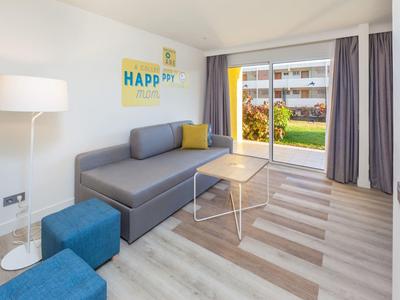 Moderner Wohnraum mit grauem Sofa, blauem Hocker, Stehlampe und großem Fenster mit Blick ins Grüne.
