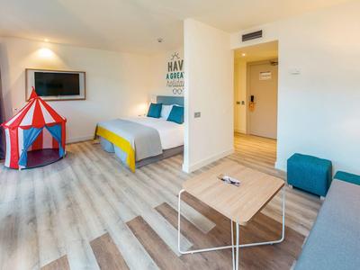 Helles Zimmer mit Holzboden, Doppelbett, Fernseher, Spielzelt und kleinem Tisch mit Sofa.