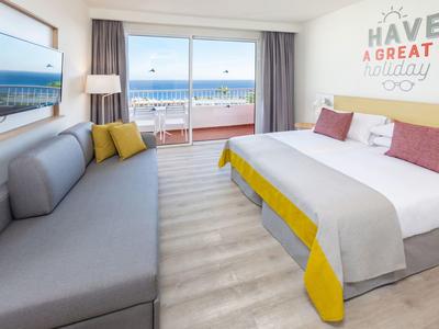 Helles Hotelzimmer mit Bett, Sofa, Meerblick und Balkon, hellgraß, gelb und weiß gestaltet.