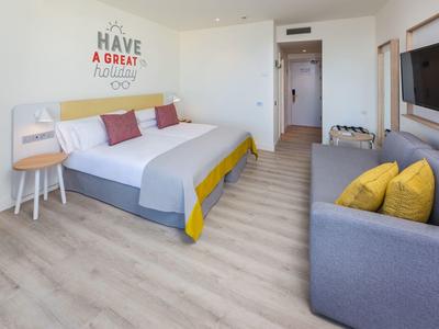 Helles, modernes Schlafzimmer mit Doppelbett, grauem Sofa und farbenfroher Wanddekoration.