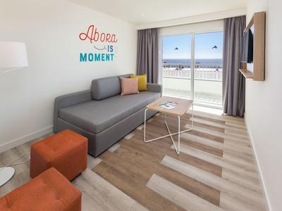Helles Wohnzimmer mit grauem Sofa, bunten Kissen, Holzfußboden und Meerblick durch großes Fenster.