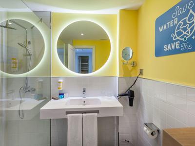 Kleine gelbe Badezimmerecke mit rundem beleuchtetem Spiegel, Waschbecken, Duschkabine und Umweltschild.