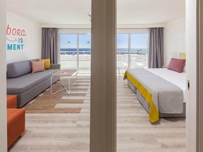 Modernes, helles Hotelzimmer mit Bett, Sofa, Teppich und großer Fensterfront, geteilt durch Wand.