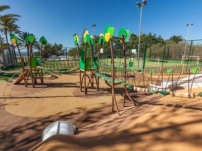 Kinder-Spielplatz mit Klettergerüst, Schaukeln, Rutsche und Tennisplatz im Hintergrund bei Sonnenschein.