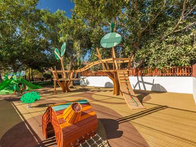 Kinderspielplatz mit Holz-Klettergerüst, Rutsche, Schaukeln und umgeben von Bäumen und Zaun.