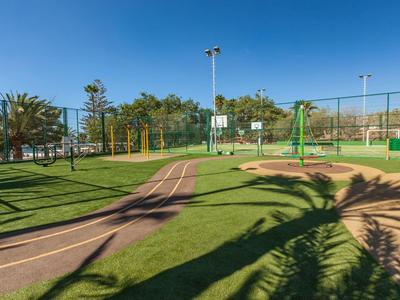 Grüner Park mit Rasenfläche, Laufbahn und Basketballkorb unter blauem Himmel.