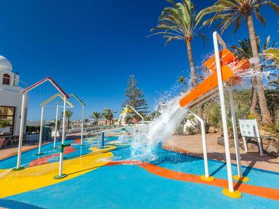 Bunter Kinderspielplatz mit Wasseranlagen, Rutschen und Palmen unter strahlend blauem Himmel.