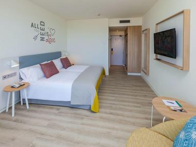 Bright hotel room with double bed, wall-mounted flat-screen TV, and cozy seating area.