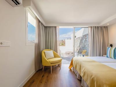 Helles Hotelzimmer mit gelbem Sessel, gelber Bettdecke und Balkon mit Bergblick.