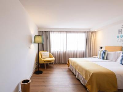 Modernes Hotelzimmer mit Doppelbett, gelber Bettdecke, Sessel und großem Fenster mit Vorhängen