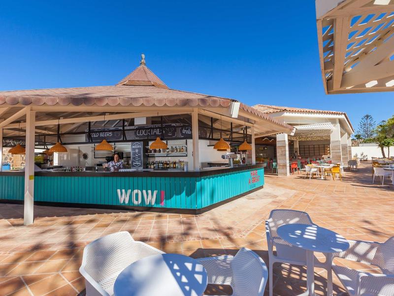 Venkovní bar s tyrkysovým pultem, bílými židlemi a slunečnou modrou oblohou.