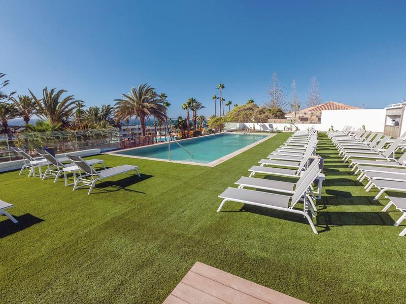 Hotelový bazén s lehátky na zelené trávě a palmami v pozadí pod modrou oblohou