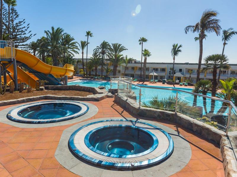 Bazénová oblast s vodní skluzavkou, dvěma vířivkami a palmami v hotelovém areálu.