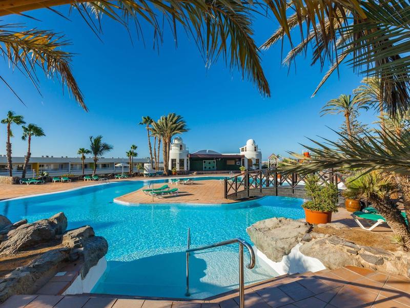 Rekreační bazén se slunečníky, palmami a jasně modrou oblohou v tropickém resortu.