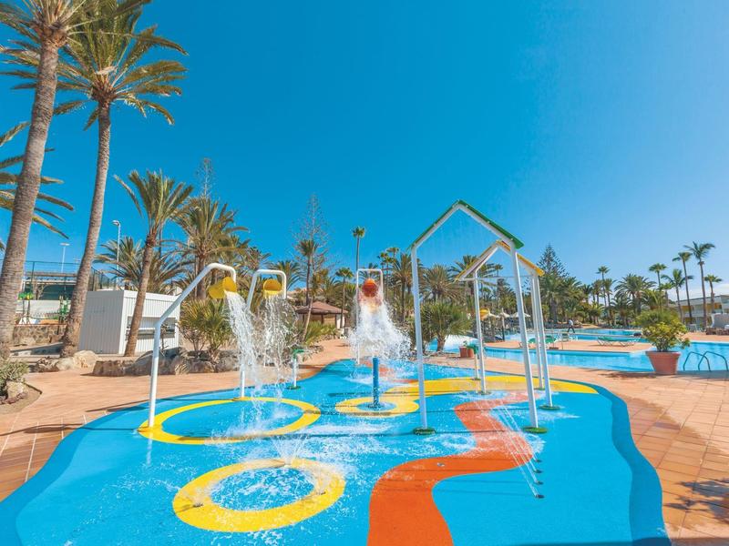 Barevná vodní herní plocha s fontánami a palmami pod jasnou oblohou.