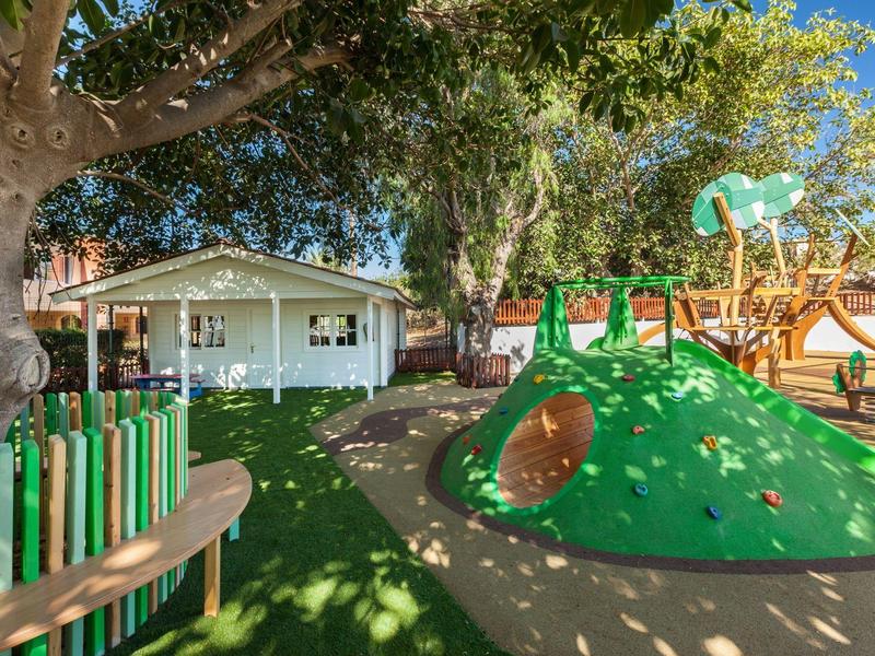 Slunné dětské hřiště s lezeckým kopcem, skluzavkou a bílým herním domkem v pozadí.