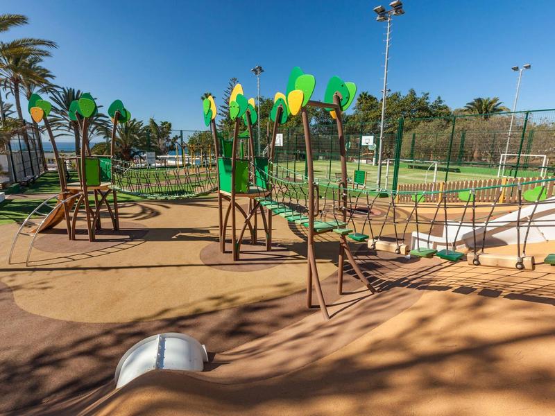 Moderní dětské hřiště s lezeckými prvky a skluzavkami v slunném hotelovém areálu.