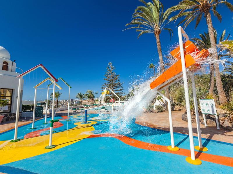 Barevné vodní hřiště s tryskajícími fontánami pod modrou oblohou a palmami