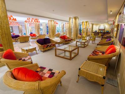 Ruime hotel lounge met rotan stoelen, rode kussens en houten tafels bij grote ramen.