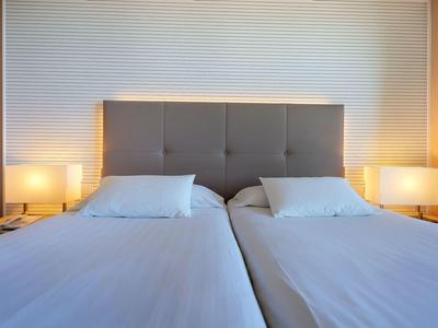 Twee eenpersoonsbedden met lichtgrijze gestoffeerde hoofdeinden en witte lakens, nachtkastjes met lampen.