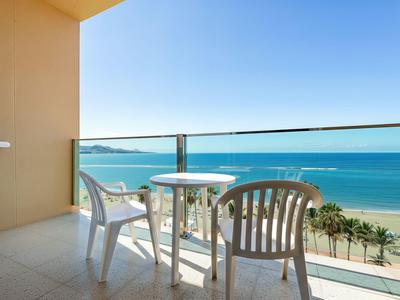 Balkon met witte tafel en stoelen met uitzicht op zee en strand bij heldere lucht.