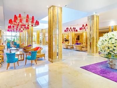 Luminoso atrio d'hotel con poltrone blu, lampade rosa e grande composizione floreale su tappeto viola.