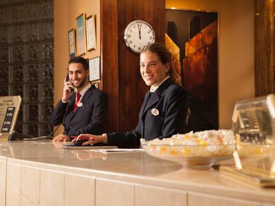 Twee hotelreceptionisten glimlachen achter een gepolijste balie met een klok aan de muur.