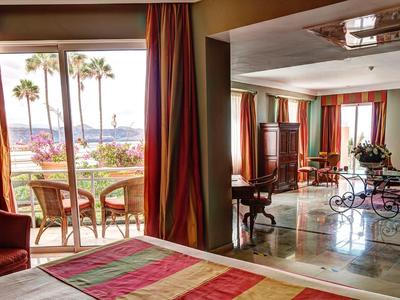 Ruime hotelkamer met bed, zithoek en groot balkon met uitzicht op palmbomen en de oceaan.