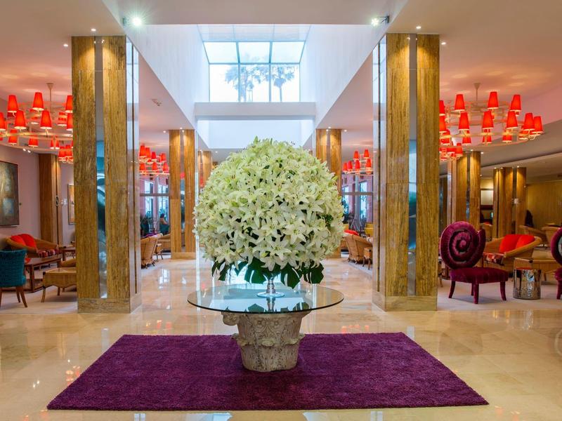 Hotel lobby met gepolijste vloeren, grote bloemdecoratie, rode kroonluchters en kleurrijke zitplaatsen.