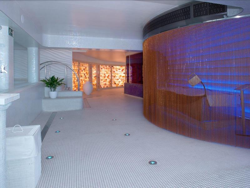 Overdekt zwembad met gebogen blauwe mozaïekmuur en zitplaatsen, witte vloer en muren, sfeerverlichting.