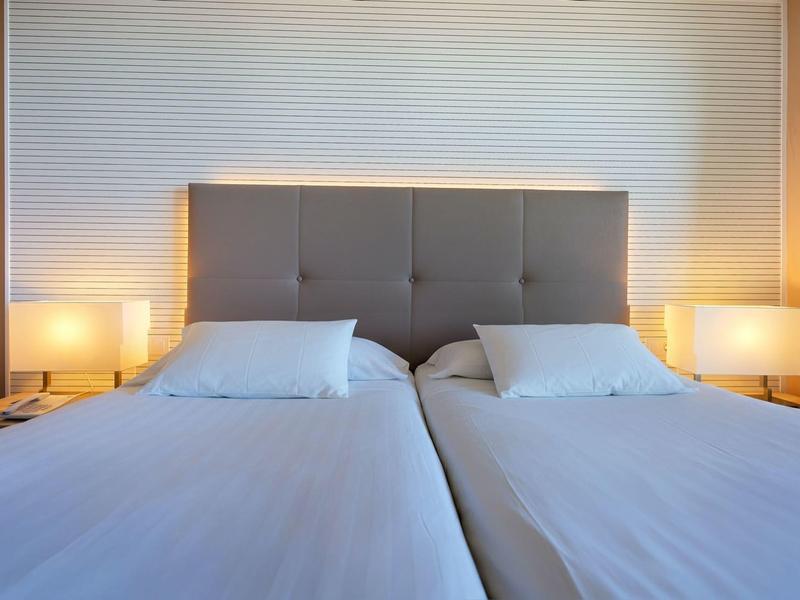 Twee eenpersoonsbedden met lichtgrijze gestoffeerde hoofdeinden en witte lakens, nachtkastjes met lampen.