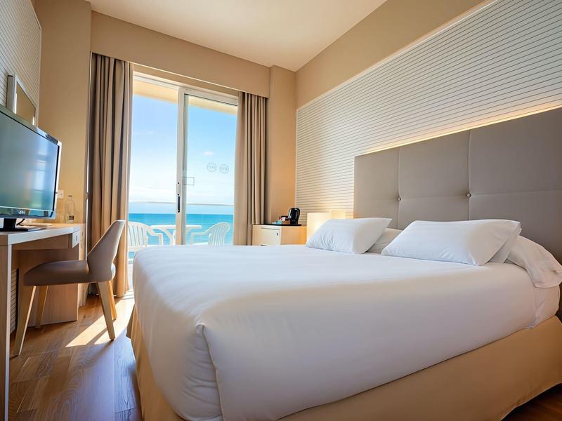 Modern hotelkamer met tweepersoonsbed, tv en uitzicht op zee door grote glazen schuifdeur.