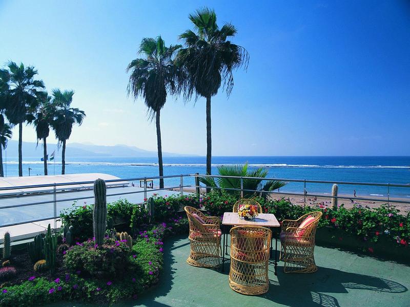 Buiten zitgedeelte met rieten stoelen en tafel bij de oceaan onder palmbomen.