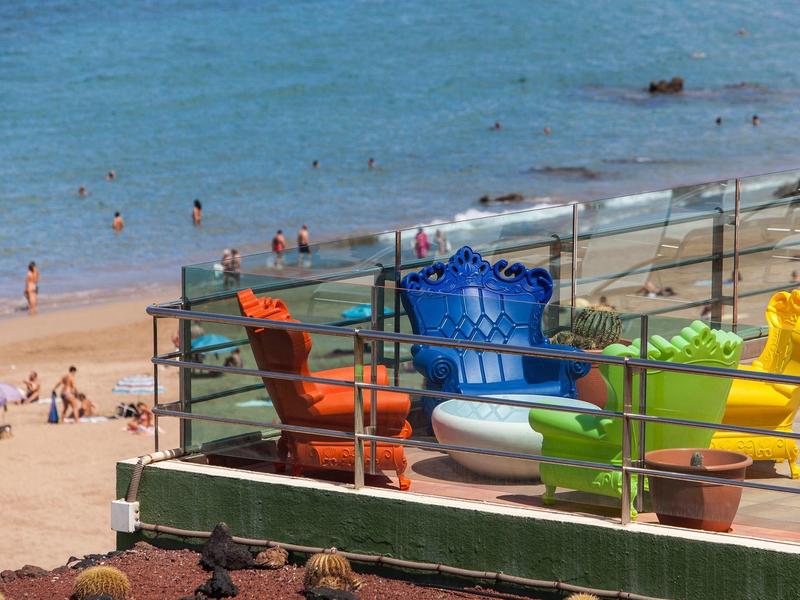 Kleurige plastic stoelen op een balkon met uitzicht op een zandstrand en oceaan met zwemmers.