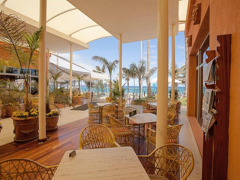 Buitenterras met tafels, stoelen en planten bij een zonnig zwembad en palmbomen.