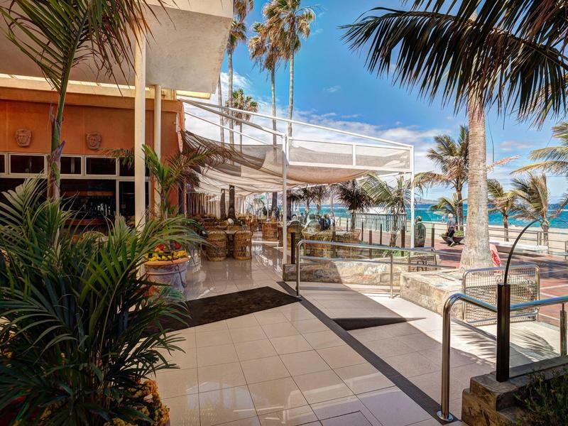 Buiten zitgedeelte van een hotel bij het strand met palmbomen en uitzicht op de oceaan.