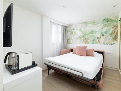 Chambre d'hôtel lumineuse avec lit double, motif végétal sur le mur et bouilloire sur armoire blanche.