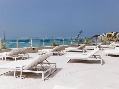 Weiße Liegestühle auf Terrasse mit Blick auf Meer und Hotelgebäude im Hintergrund.
