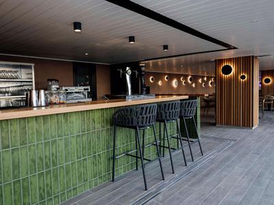 Moderner Hotelbarbereich mit grün gekacheltem Tresen, hohen Stühlen und holzvertäfeltem Hintergrund.