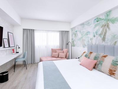 Helles Hotelzimmer mit Doppelbett, farbigen Kissen, Lounge und tropischer Wandgestaltung.