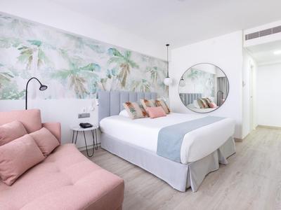 Helles Hotelzimmer mit Doppelbett, rosa Couch und Wand mit tropischem Muster.