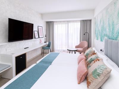 Helles Hotelzimmer mit Doppelbett, farbigen Kissen, Schreibtisch und Fernseher.