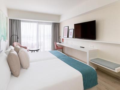 Helles Hotelzimmer mit großem Bett, Fernseher an der Wand und Balkon mit Blick nach draußen.
