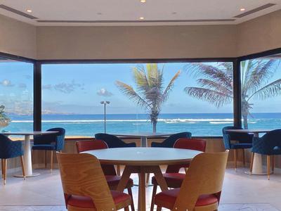 Comedor moderno con vista al mar y palmeras, mesas y sillas a la luz natural.