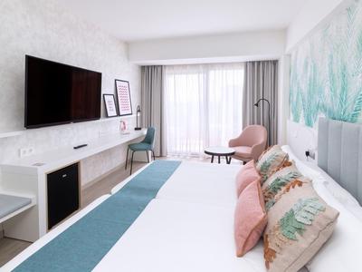 Chambre d'hôtel lumineuse avec grand lit, oreillers colorés, TV murale et balcon