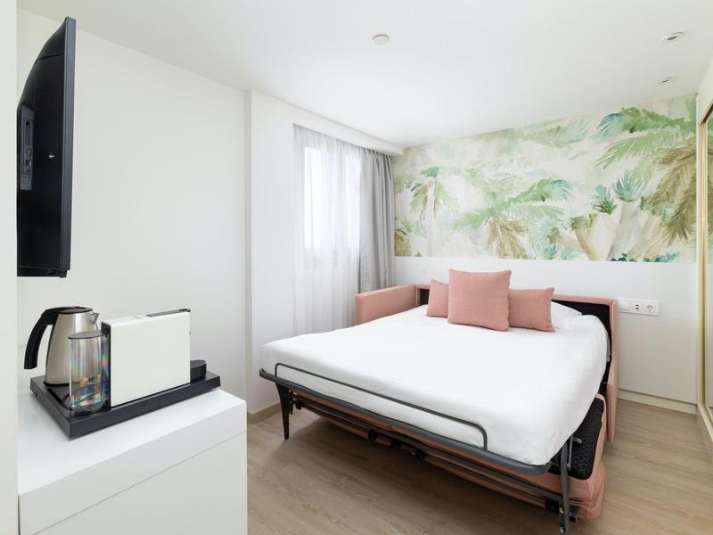 Chambre d'hôtel lumineuse avec lit double, motif végétal sur le mur et bouilloire sur armoire blanche.