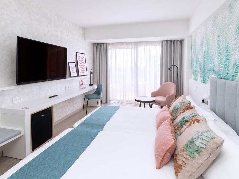 Chambre d'hôtel lumineuse avec grand lit, oreillers colorés, TV murale et balcon