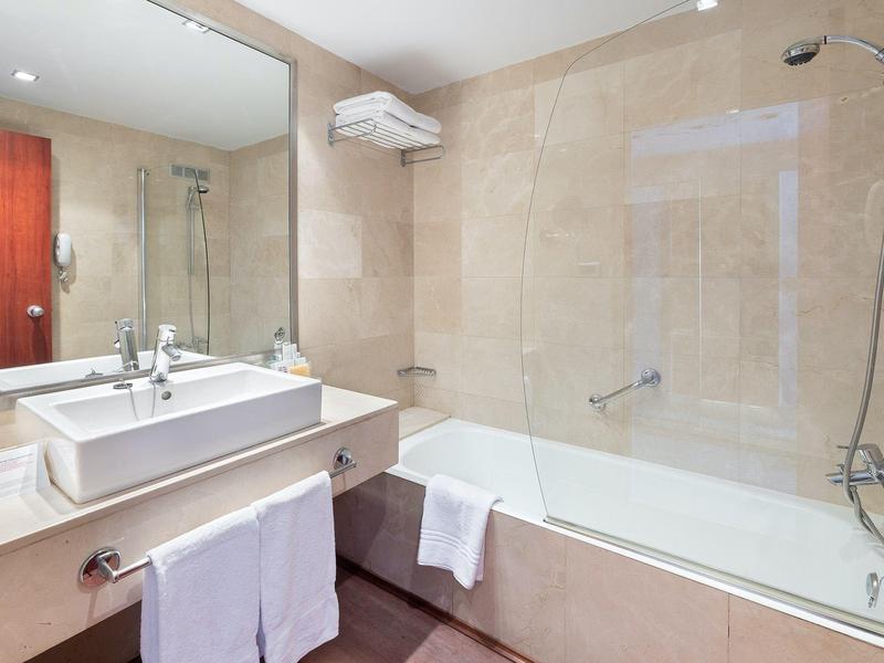 Salle de bain lumineuse avec baignoire, douche, grand miroir et lavabo avec serviettes.