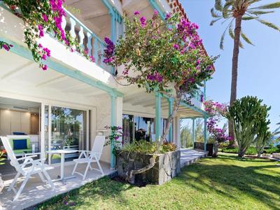 Terrasse d'hôtel avec plantes en fleurs, chaises et tables blanches, entourée de pelouse verte et de palmiers.